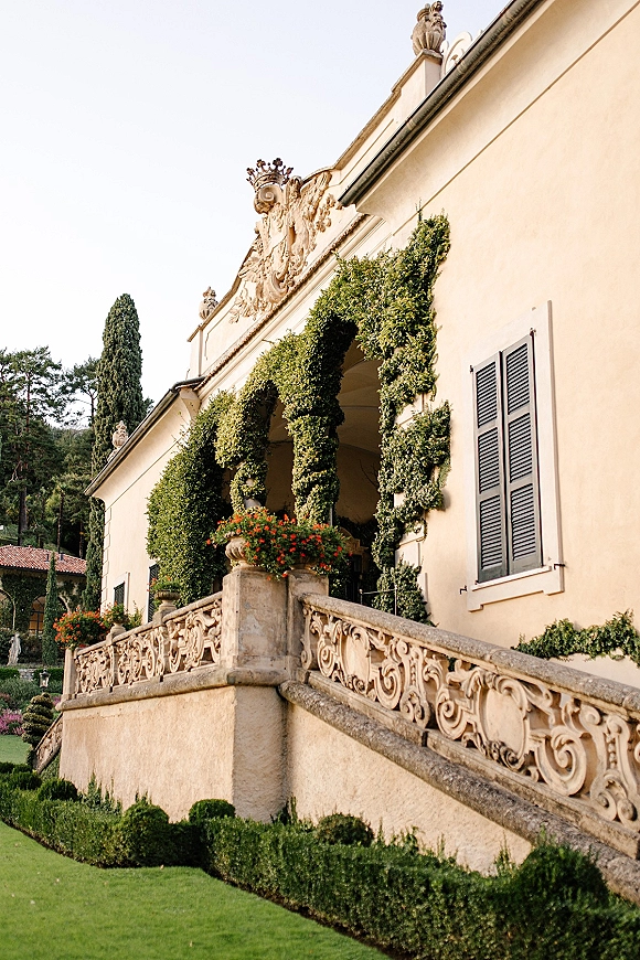 Wedding venue exterior with climbing ivy and stone balustrade, cream stucco villa and black shutters above manicured garden lawn on hillside