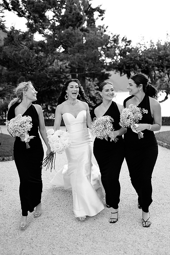 Bridesmaids photo of a bride with bridesmaids walking and laughing on a garden path, holding baby’s breath bouquets with ribbon wraps near water