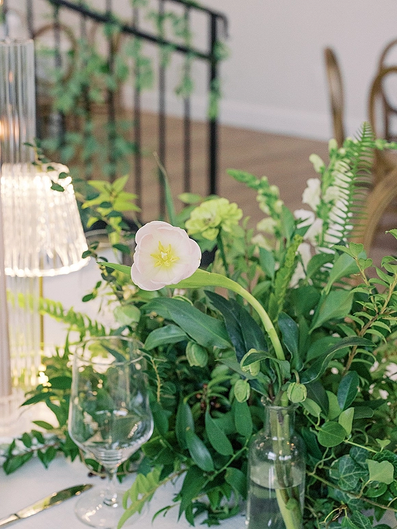 Reception centerpiece with greenery wedding centerpiece in a glass vase, single white flower, wine glass and lamp on black metal stand nearby