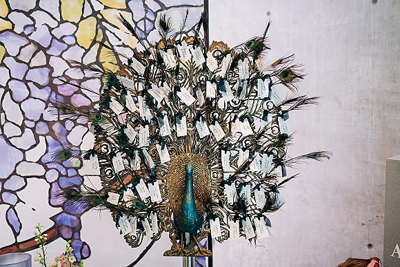 Wedding escort display with peacock escort cards hanging from ribbons on an ornate metal stand before a stained glass wall