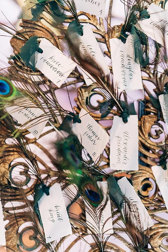 Escort cards with calligraphy escort cards on torn-edge paper, tied with green ribbons and peacock feather accents in an ornate gold frame display