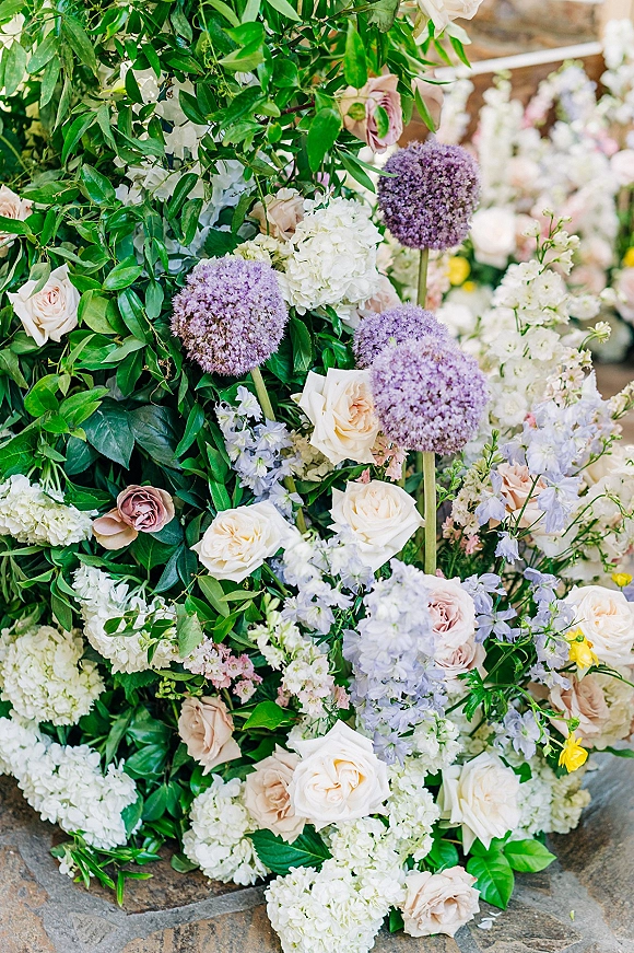 Wedding floral arrangement of pastel wedding flowers with roses, hydrangeas, purple allium and greenery on a stone floor, blurred blooms behind
