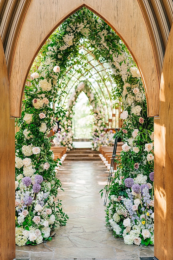 Ceremony aisle decor with a floral ceremony entrance featuring a lush arch of white blooms and blush roses at wooden chapel doors along a stone aisle