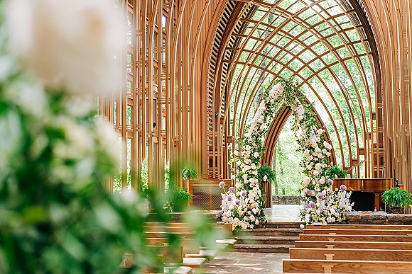 Ceremony setup with a floral ceremony arch of white and blush blooms, wooden pews and aisle steps beneath a vaulted glass chapel ceiling