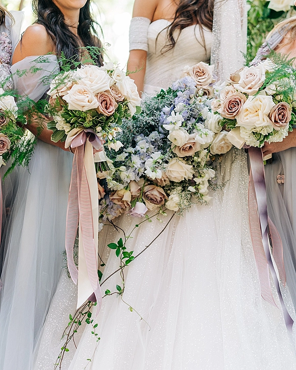 Bridesmaid bouquets in a neutral rose bouquet palette with roses, ranunculus and delphinium, trailing greenery and ribbon streamers outdoors