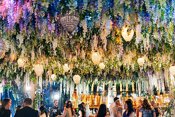 Reception dance floor with wedding dance floor lighting under hanging floral ceiling decor, crystal chandeliers, and greenery as guests dance by glass walls