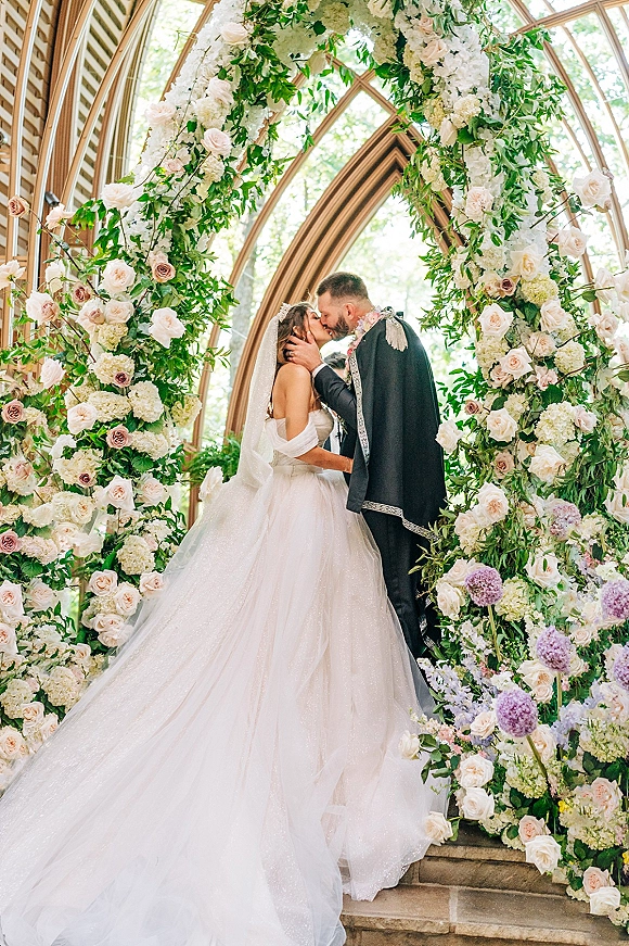 Wedding kiss portrait of bride and groom kissing beneath a rose and hydrangea floral arch, framed by glass conservatory windows and stone steps