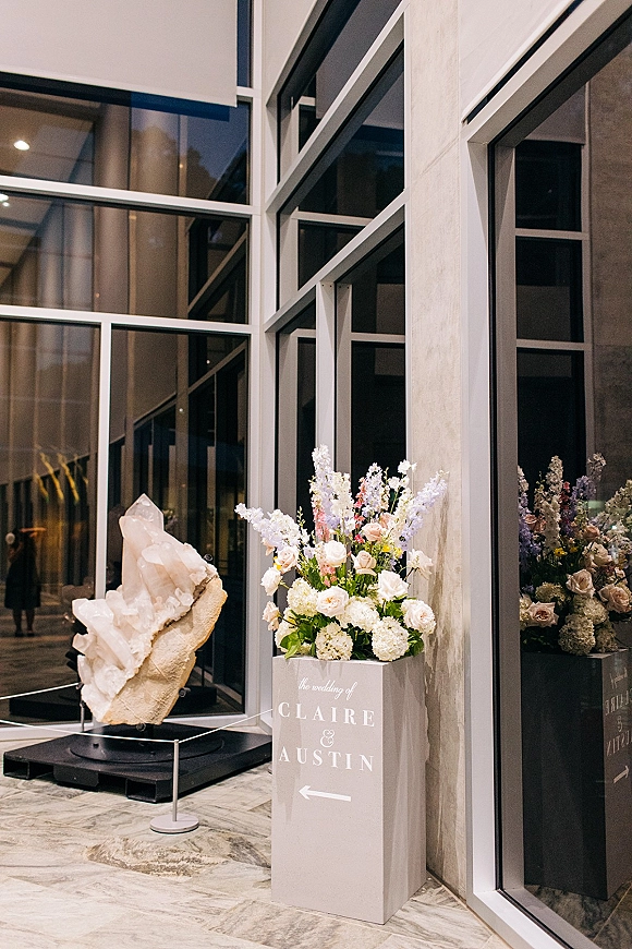 Wedding welcome sign with flowers on a white pedestal and arrow in a modern lobby with glass windows and sculpture display