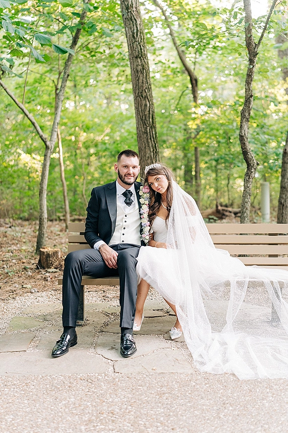 Couple portrait of bride and groom sitting on bench in woodland, her long veil and tiara, holding a pink bouquet beside him in tuxedo