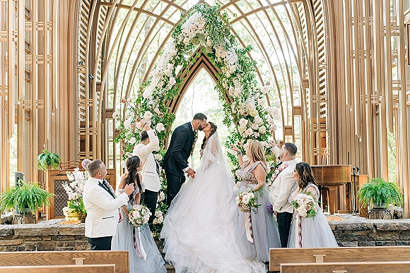 Wedding kiss as bride and groom embrace beneath a floral arch with greenery and blush roses inside an arched-window chapel, veil flowing