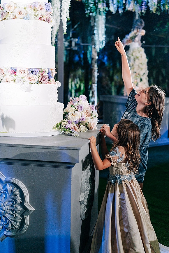 Wedding cake four tier wedding cake with white buttercream, pastel roses and lavender topper on a table under hanging florals in a dark venue
