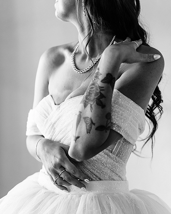 Bridal portrait in black and white of a bride in an off the shoulder wedding dress adjusting her beaded necklace against a plain wall