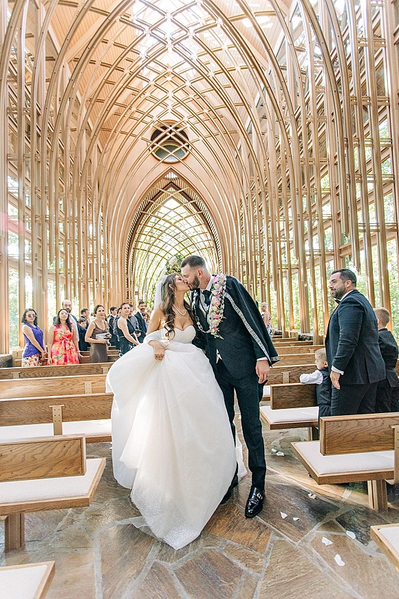 Wedding kiss as newlyweds share a ceremony kiss moment in a chapel aisle, bride in strapless ball gown and veil, guests in pews