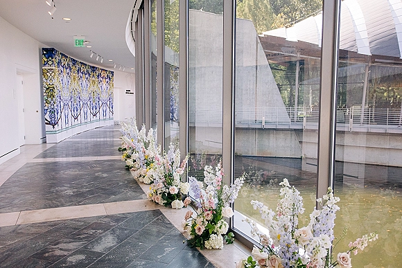 Ceremony aisle flowers in a lush ground arrangement of white and blush roses, hydrangea and delphinium in a modern glass hallway