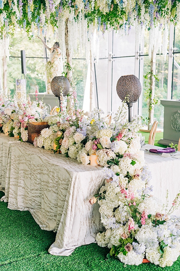 Head table decor with a floral garland runner of hydrangeas and roses, taper and pillar candles, purple napkins, beneath hanging florals by glass walls