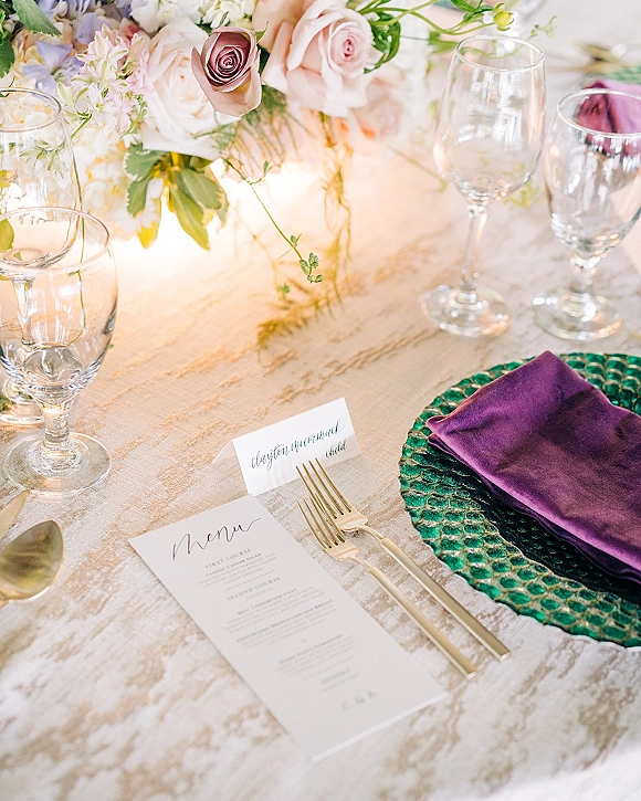 Reception tablescape with wedding place setting, rose centerpiece, velvet napkin, green glass charger, gold flatware, and candlelight on linen tablecloth