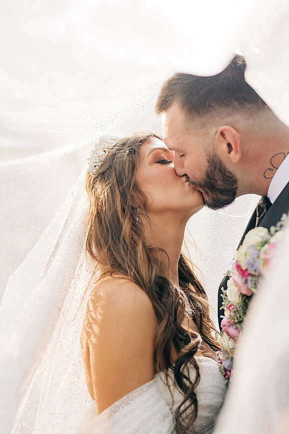 Wedding kiss as bride and groom kiss under her veil, tiara and drop earrings visible, holding a pastel bouquet in soft sky light