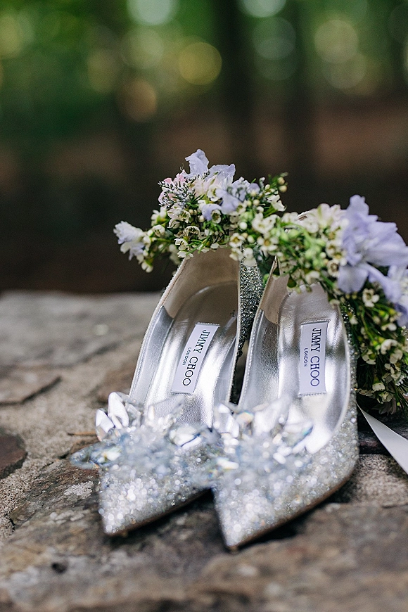 Bridal shoes flatlay of silver glitter heels with crystal bow on a stone surface, styled with a floral crown and ribbon by greenery
