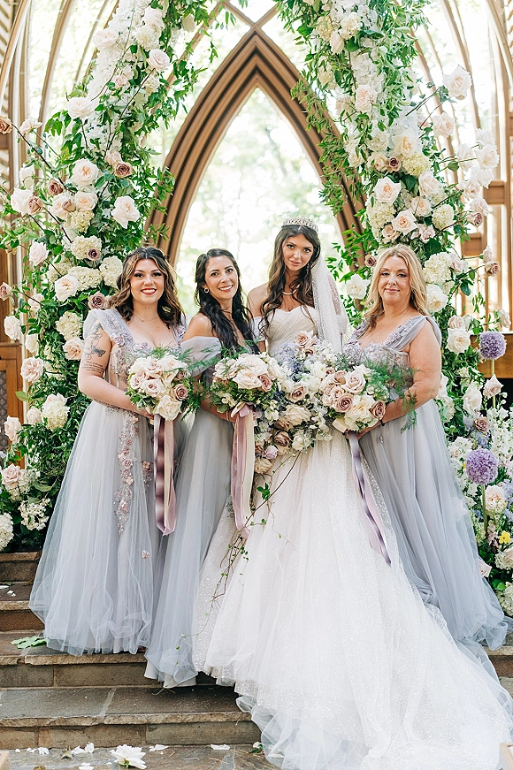 Bride with bridesmaids group photo on stone steps, bride in veil and tiara holding bouquets with ribbon tails by chapel windows and floral arch