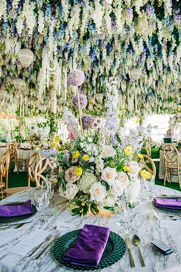 Reception tablescape with wedding table centerpiece of white and lavender blooms, purple napkins and goblets beneath hanging florals in tent canopy
