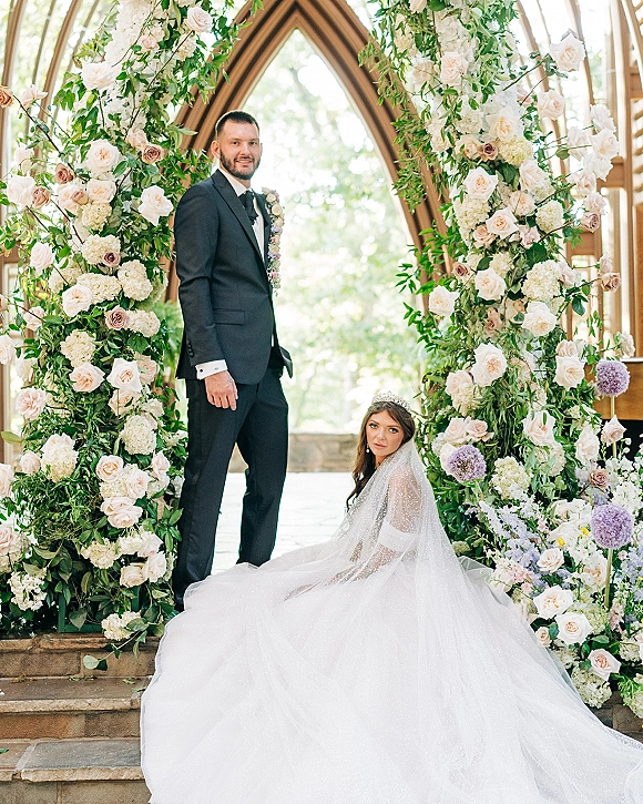 Couple portrait of bride seated on stone steps in cathedral veil and tiara as groom in tux stands beside floral arch by arched windows