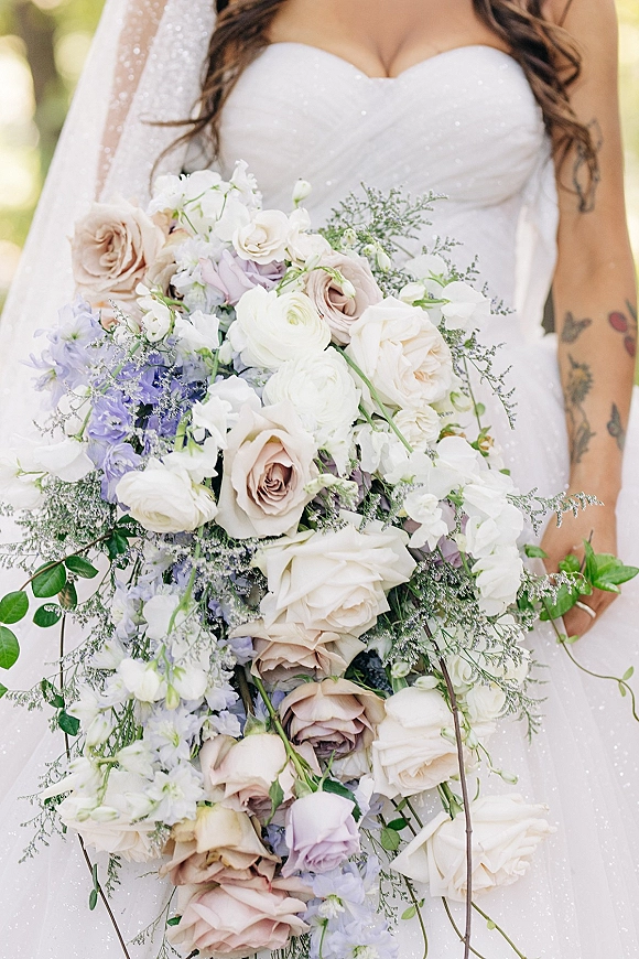Bridal bouquet with cascading bridal bouquet of ivory roses, ranunculus and lavender blooms in bride’s tattooed hands, soft greenery bokeh behind