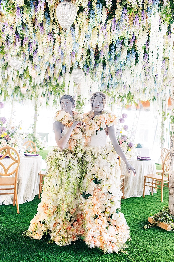 Bride and groom portrait in flower dress wedding attire beneath hanging florals and chandeliers inside a white tent reception with round tables