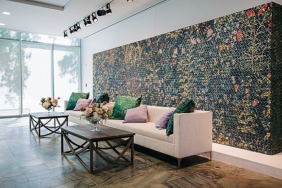 Wedding lounge seating with a white sofa, wood coffee tables, and floral centerpieces set before a floral wall and large windows