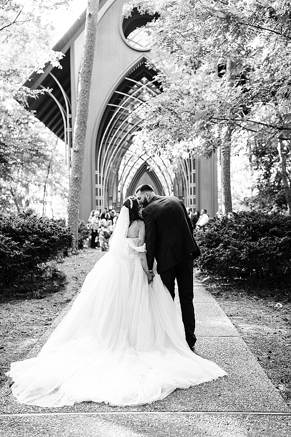 Wedding couple portrait in black and white, bride and groom from behind holding hands on a chapel walkway with guests and arched backdrop