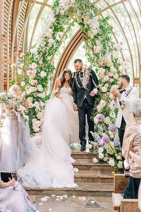 Wedding recessional as bride and groom walk the aisle holding hands, petals falling beneath a rose arch in a glass chapel with guests clapping