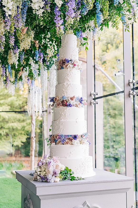 Wedding cake with tiered white buttercream and cascading fresh flowers on a pedestal beneath hanging florals by glass windows, garden view