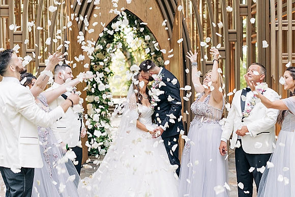 Wedding kiss portrait as bride and groom hold hands in chapel doorway, rose petal exit confetti flying, veil trailing in light