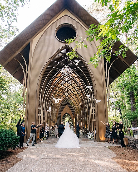 Wedding recessional as the bride in a gown and long veil exits an arched wooden chapel while guests release white doves onto a stone walkway