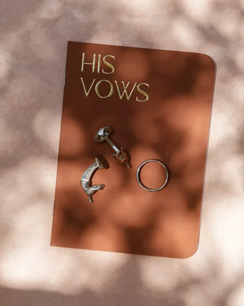 Wedding vow book with gold foil lettering beside a wedding ring and cufflinks on beige fabric in soft shadows