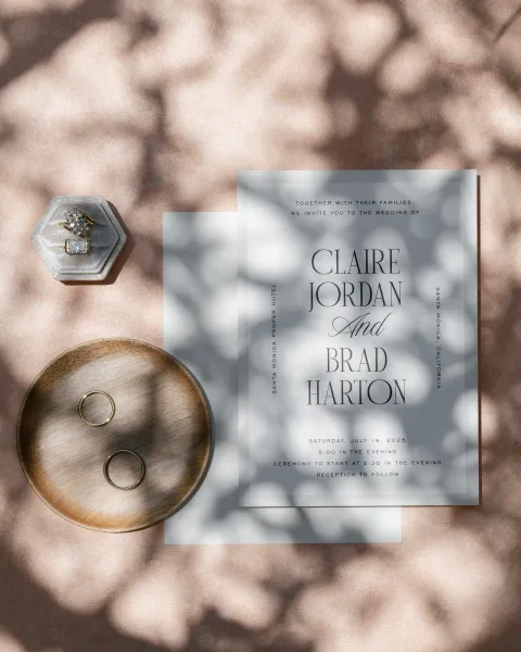 Invitation suite wedding invitation flat lay with vellum overlay, envelope, and rings beside a ring box on pink fabric in dappled light