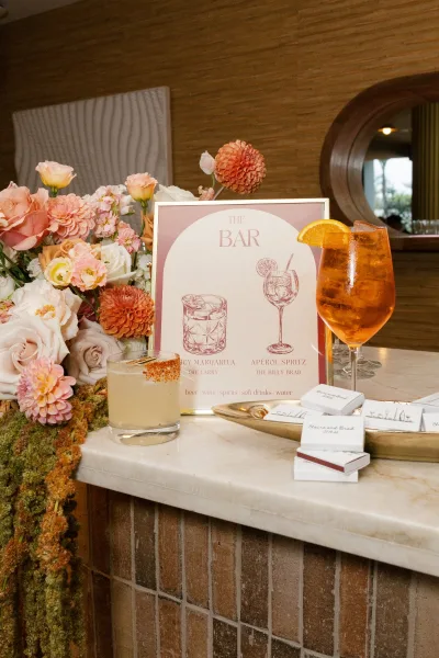 Wedding bar sign and signature cocktail sign in a frame beside rose and dahlia florals on a marble countertop with orange-garnished glasses
