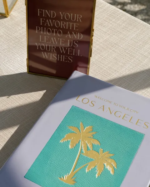 Wedding welcome sign in a gold frame, acrylic card with gold foil lettering on linen table, sunlight casting shadows on pavement