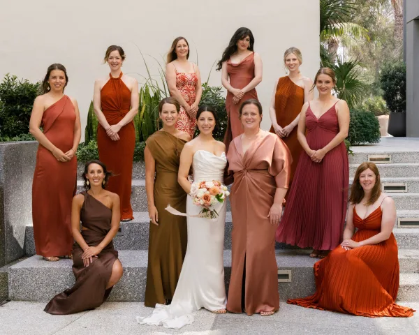 Bride with bridesmaids in terracotta dresses posing on stone steps, bride in strapless gown holding bouquet, palm greenery behind