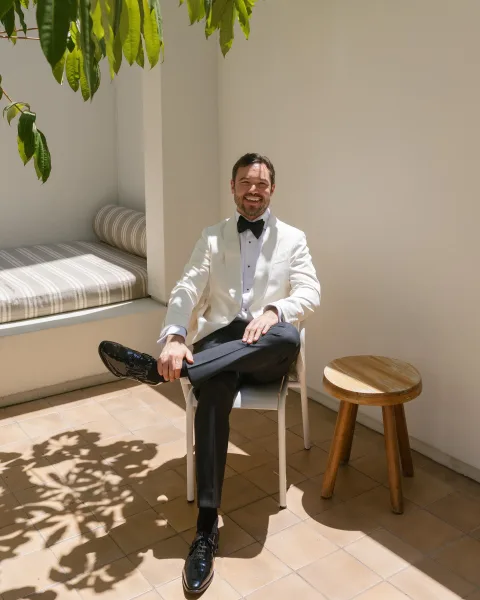 Groom portrait in a white tuxedo jacket and black bow tie, seated on a chair by a stucco wall with sunlit shadows on tiled floor