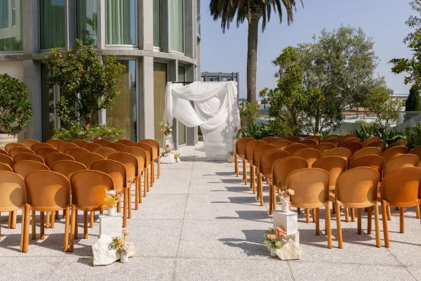 Ceremony setup with wood chairs and fabric-draped arch lined with peach and white florals on a modern outdoor terrace by a palm tree