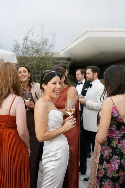 Wedding cocktail hour as bride holding champagne chats with guests on an outdoor patio by a modern venue, glasses raised in candid mingling