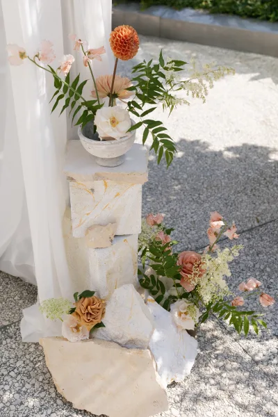 Ceremony florals with wedding aisle florals in a ceramic bowl, featuring peach blooms, white roses, and greenery on a stone pedestal by hedge