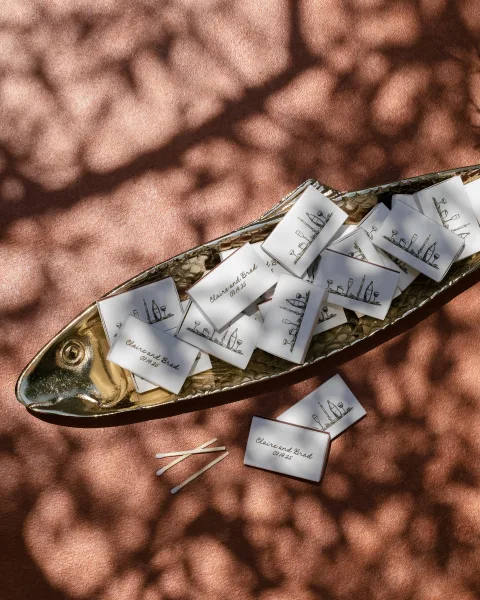 Wedding matchboxes with personalized labels arranged on a gold tray, matches nearby on a textured pink surface with dappled shadows