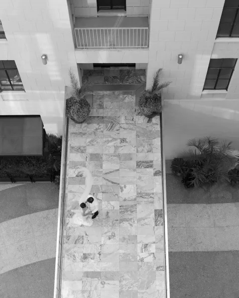 Couple portrait from above of bride in a long veil and groom in suit embracing on a stone tile courtyard beside potted palms