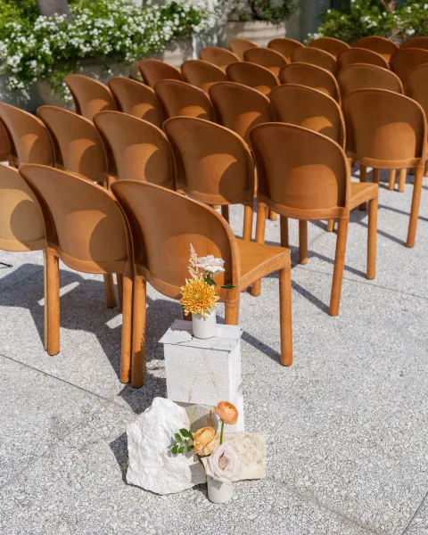 Ceremony seating with tan leather chairs lined on gravel, bud vases and white pedestals marking the aisle with roses and orange blooms