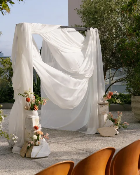 Wedding ceremony arch with white drapery and floral greenery accents on stone plinths, set on a terrace with ocean view beyond