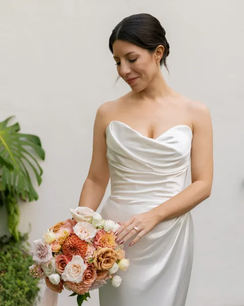 Bridal portrait of a bride in a strapless satin wedding dress with draped neckline, holding rose and dahlia bouquet by greenery and monstera leaves