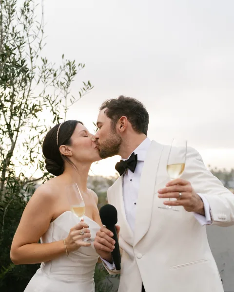 Wedding kiss as newlyweds toasting champagne, bride in strapless dress and pearl headband, groom in tux, outdoors with trees and sky