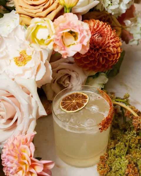 Wedding signature cocktail with a citrus garnish in a rocks glass, chili salt rim and ice, styled beside peach blush flowers on a neutral tabletop