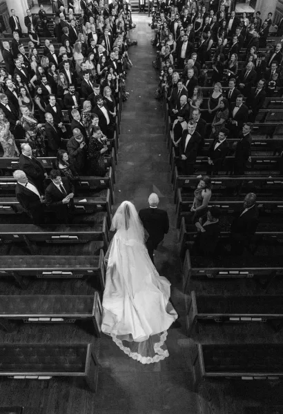 Ceremony processional with the bride walking down the aisle, her veil sweeping past wood pews in the church sanctuary interior.
